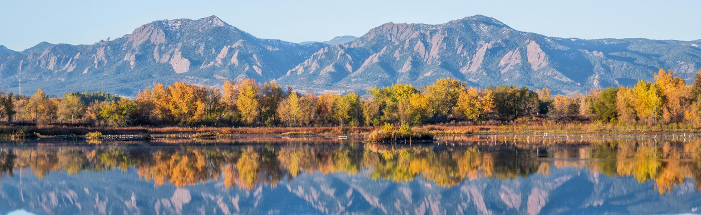 The Boulder Flatirons in the background with a clear lake in the foreground and fall trees.
