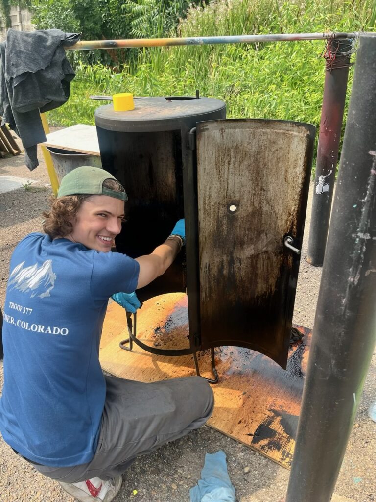 A young man smiles while cleaning out a metal meat smoker/BBQ. 