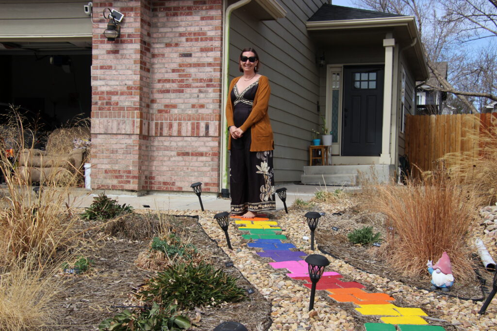A woman standing in front of her home, with a rainbow colored path and garden in front of her.