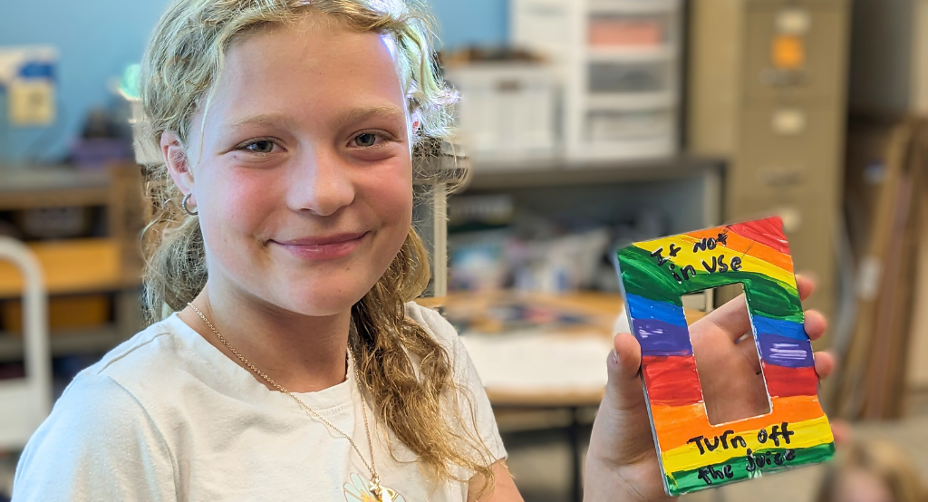 Renew Our Schools student holding a light switch plate with the words "If not in use, turn off the juice.