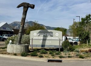 A large sculpture of a hammer made of recycled materials stands in front of a sign for Eco-Cycle and Resource Central on Arapahoe Road in Boulder, Colorado.