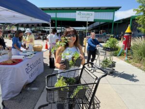 A person holds two potted plants, smiling at the camera, with a shopping cart full of plants and a Resource Central logo in the background at an outdoor market.