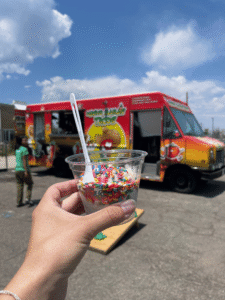 A hand holds a clear plastic cup filled with ice cream and rainbow sprinkles, with a white spoon. In the background, a colorful food truck under a bright blue sky with clouds.