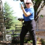 A person in a blue shirt holds a tablet and a blue catch cup while standing on a lawn with several blue catch cups on the grass in front of a house during a Slow the Flow sprinkler evaluation with a bench in the background.