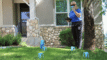 A person in a blue shirt holds a tablet and a blue catch cup while standing on a lawn with several blue catch cups on the grass in front of a house during a Slow the Flow sprinkler evaluation.