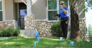 A person in a blue shirt holds a tablet and a blue catch cup while standing on a lawn with several blue catch cups on the grass in front of a house during a Slow the Flow sprinkler evaluation.