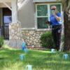 A person in a blue shirt holds a tablet and a blue catch cup while standing on a lawn with several blue catch cups on the grass in front of a house during a Slow the Flow sprinkler evaluation.