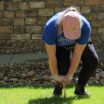 A person in a blue shirt kneeling on a lawn with a soil sampler probe the grass in front of a house during a Slow the Flow sprinkler evaluation.
