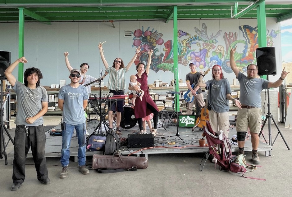 A band holding instruments stands on a stage with a colorful mural behind them. One person holds a baby, and several band members have their arms raised in the air.