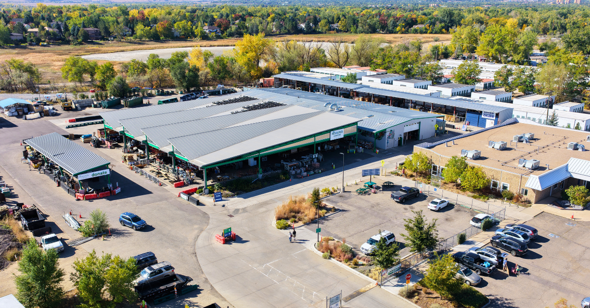 Aerial view of Resource Central's nonprofit home improvement center with multiple metal-roof buildings, drive-through donation lane, loading areas, parked trucks and cars, and a paved yard surrounded by trees.